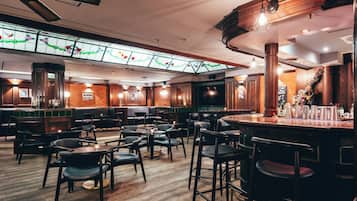 Un espace bar avec des tabourets de bar en bois, un plafond haut avec des poutres apparentes et un vitrail au-dessus.
