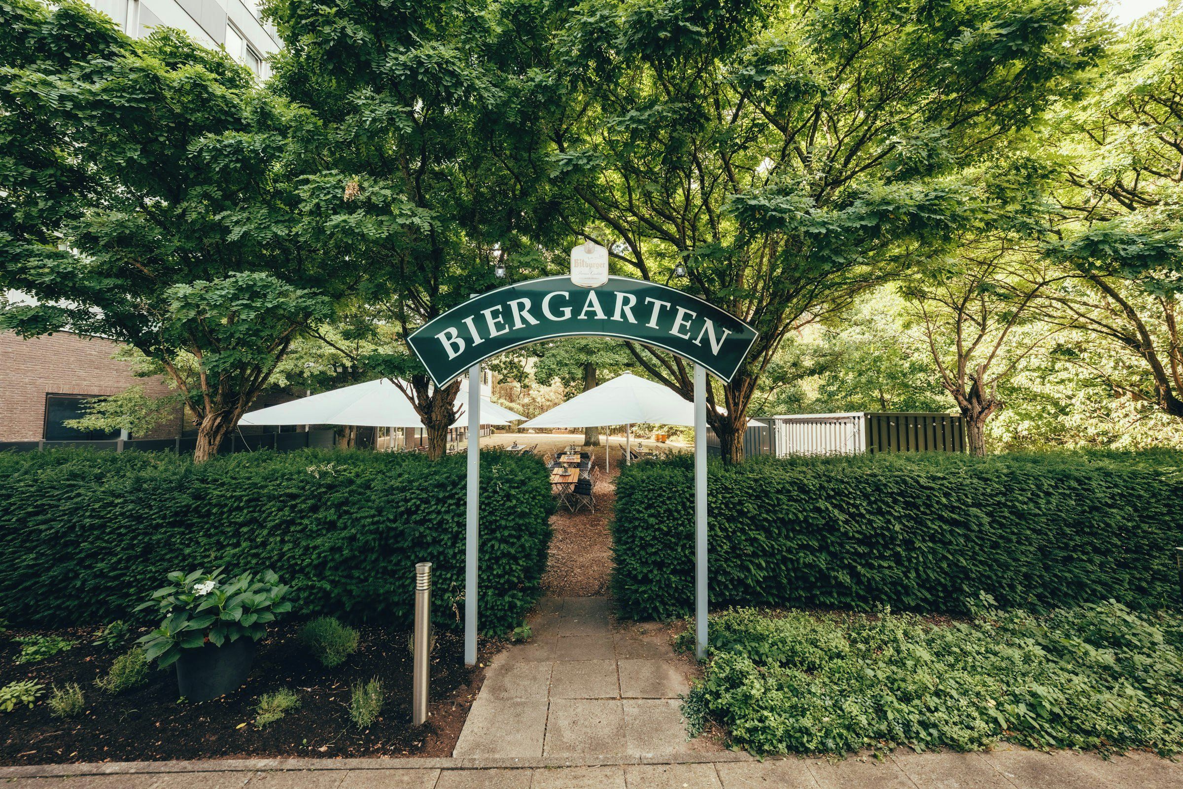 A pathway leading to a sign reading 'Biergarten' with umbrellas and greenery.