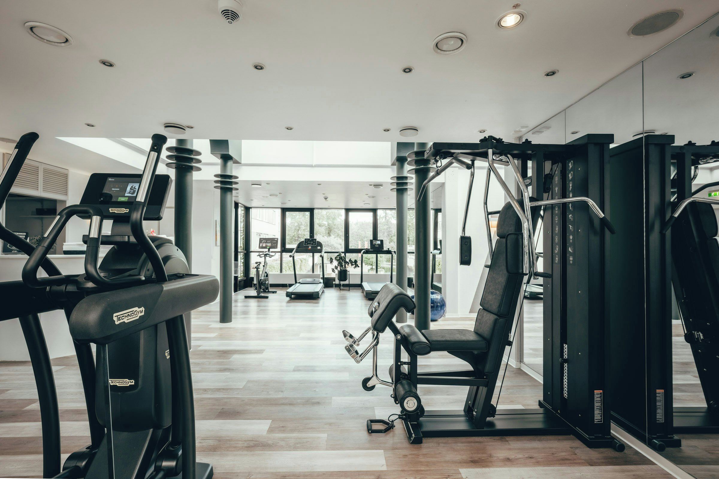 Un gimnasio con máquinas de cardio, equipo de levantamiento de pesas y un amplio ventanal que proporciona luz natural.