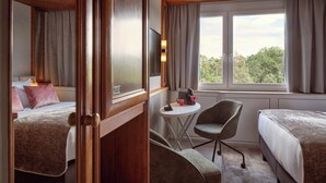 A hotel room with two beds, a small round table with a chair, a TV, and a window with curtains. - Leonardo Frankfurt City South (Frankfurt)