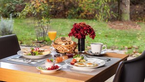 Una mesa de desayuno al aire libre con platos de comida, una cesta de pan, un florero y una taza de café.