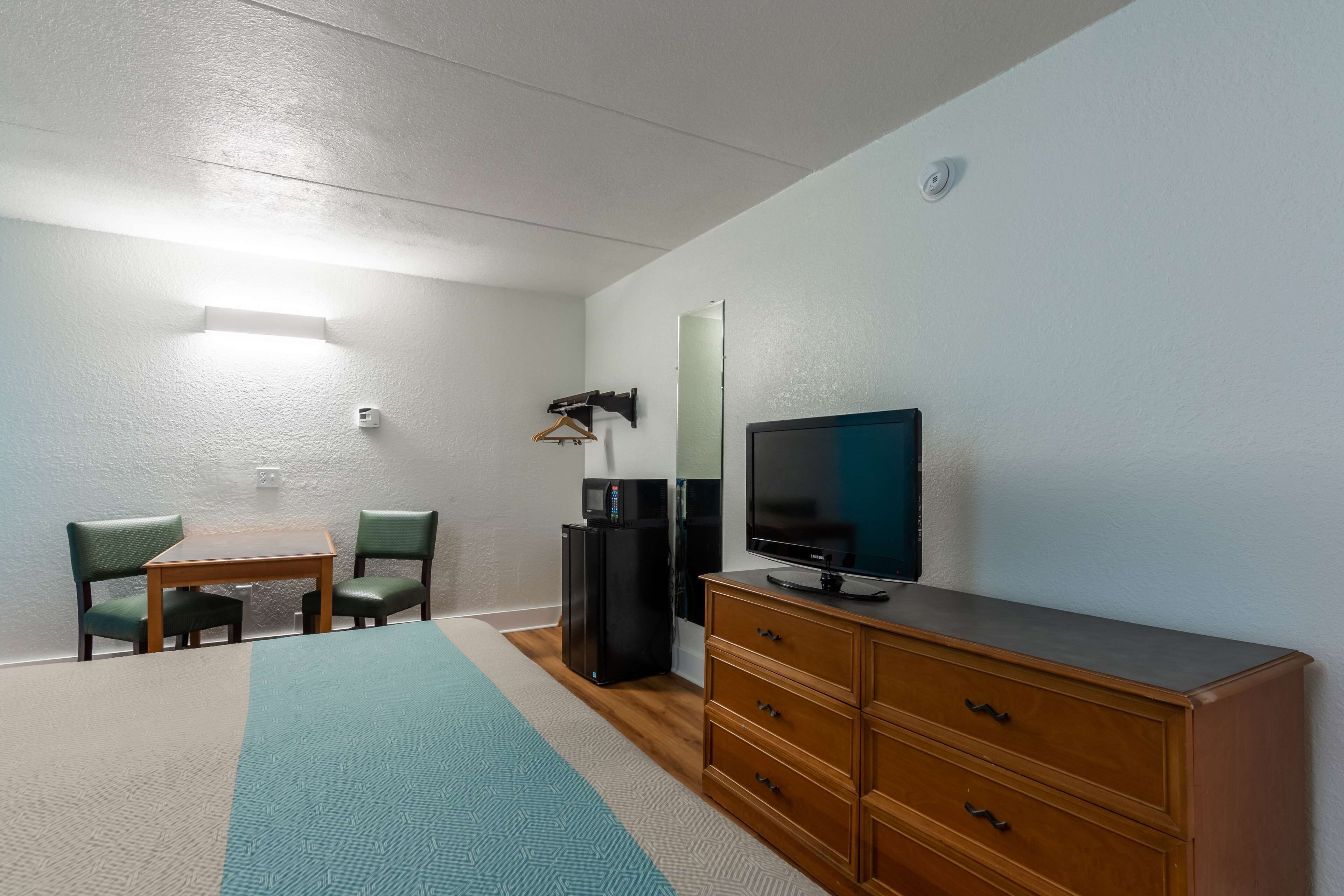Een kamer met een houten dressoir, een flatscreen-televisie, een klein tafeltje met twee stoelen, een koelkast en een wandlamp.