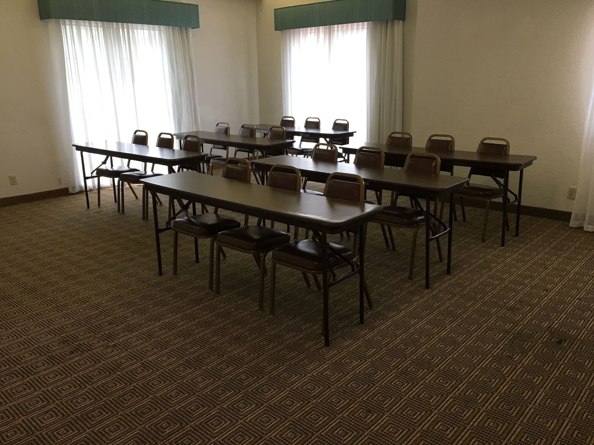 Un salón de conferencias con varias mesas y sillas, ventanas amplias con cortinas y una alfombra estampada.