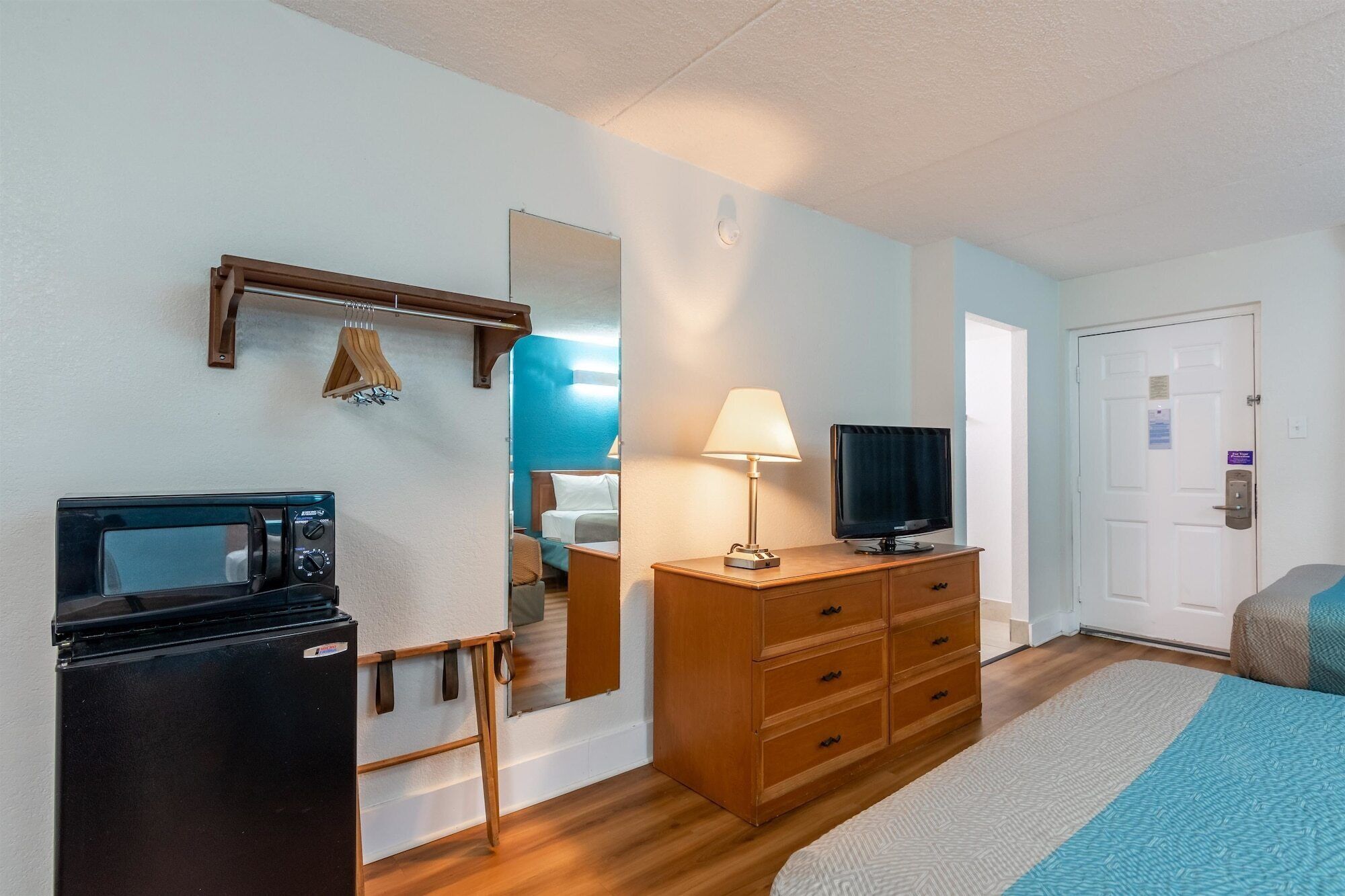 A compact hotel room with a bed, a TV, a wooden dresser, a microwave, and a small table with a lamp.
