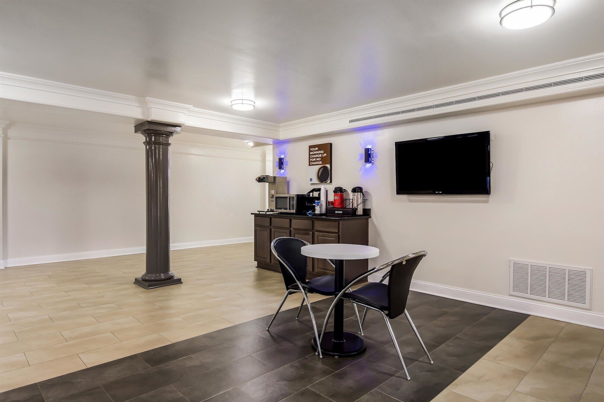 A modern, well-lit room with a dark floor, a round table, two chairs, a television, and a kitchenette area with a microwave and coffee maker.