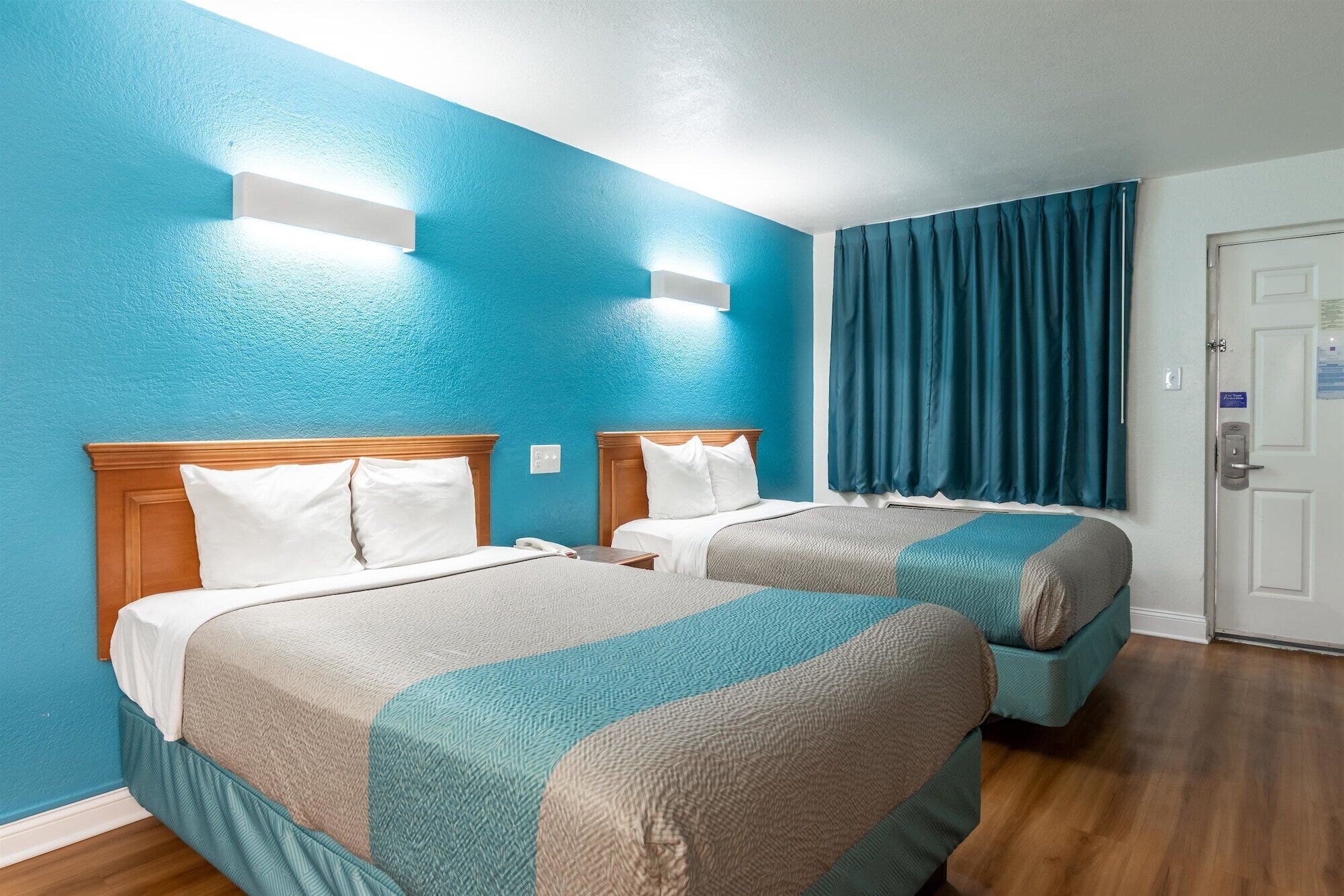 Habitación de hotel con dos camas, cabecero de madera, pared azul, ventana con cortinas y puerta con cerradura.