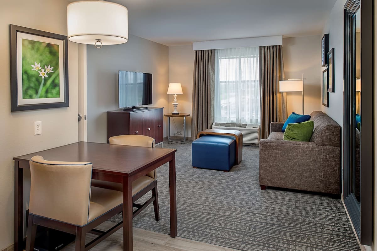 A hotel room with a dining table, a sofa, a TV, a lamp, and a framed picture on the wall.