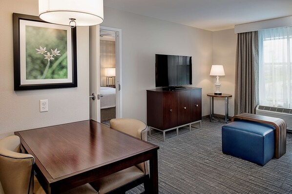 Habitación de hotel con un escritorio de madera, televisor, un reposapiés azul, un cuadro en la pared y una ventana con cortinas.