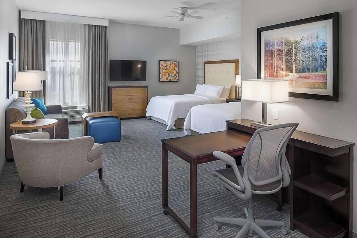 A hotel room with two beds, a desk, a chair, a TV, a painting, and a ceiling fan.