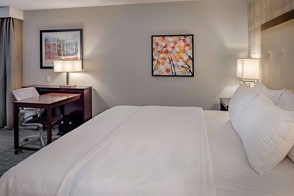 A hotel room with a large bed, a desk with a chair, two bedside lamps, and two framed artworks on the walls.