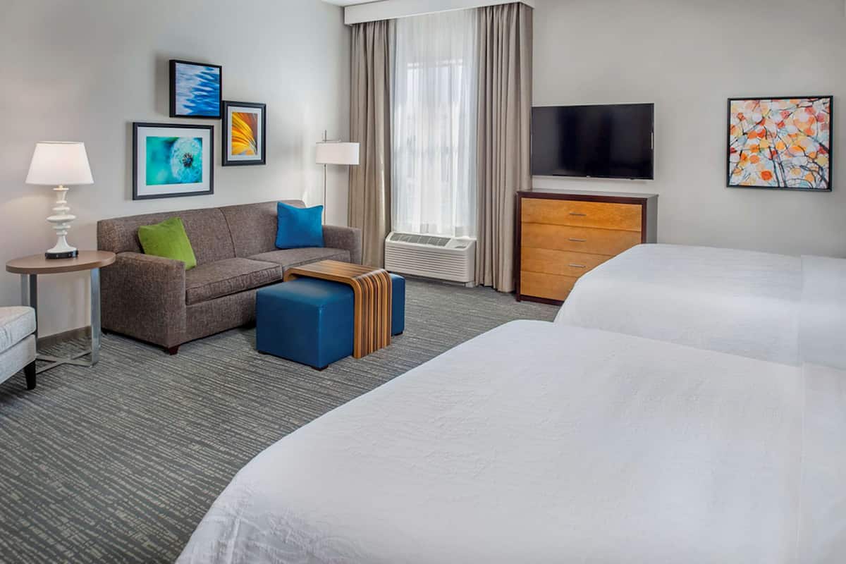 A hotel room with a sofa, two ottomans, a TV, a desk, and a bed.