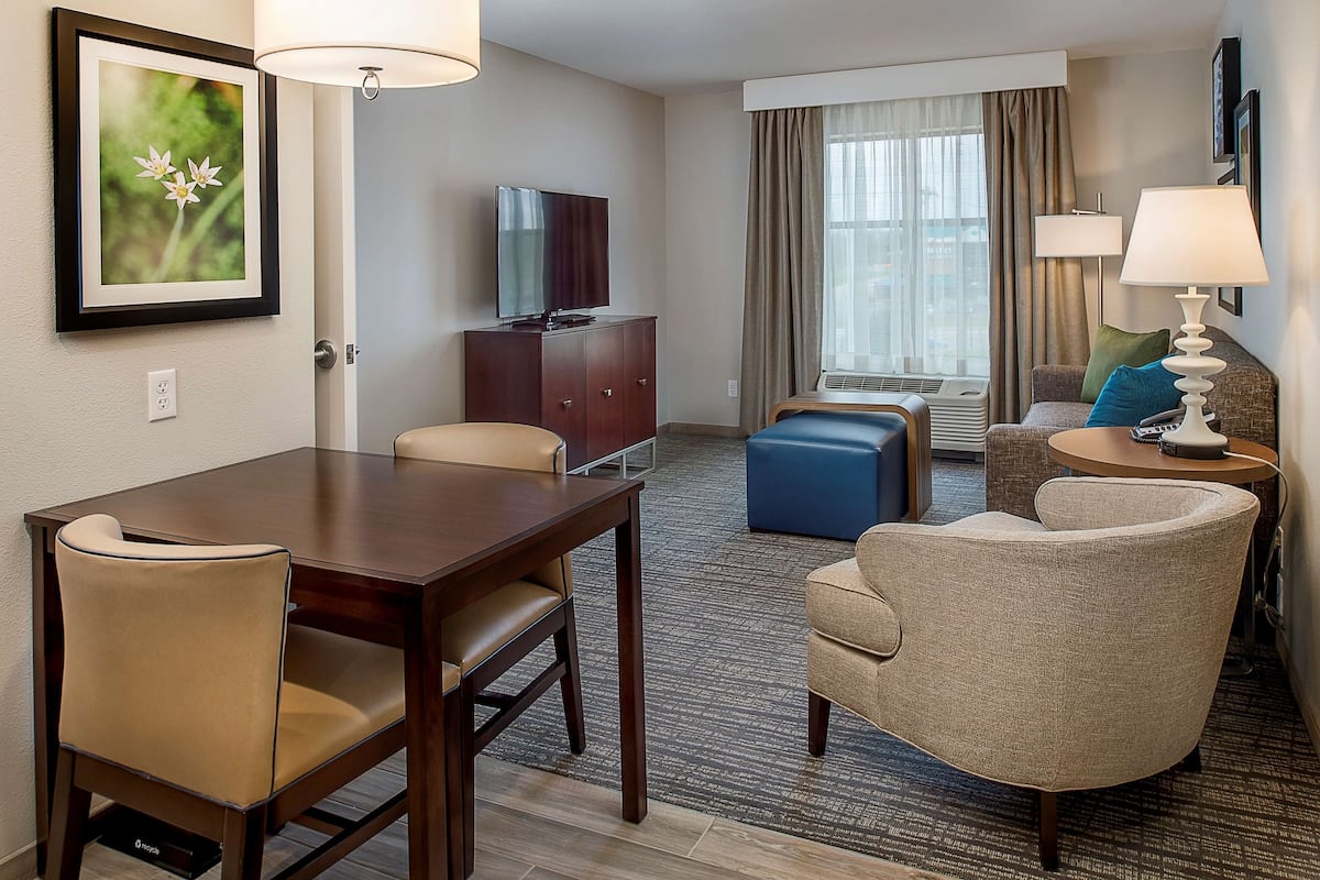 A hotel room with a dining table, two chairs, a sofa, a TV, a lamp, and a window with curtains.