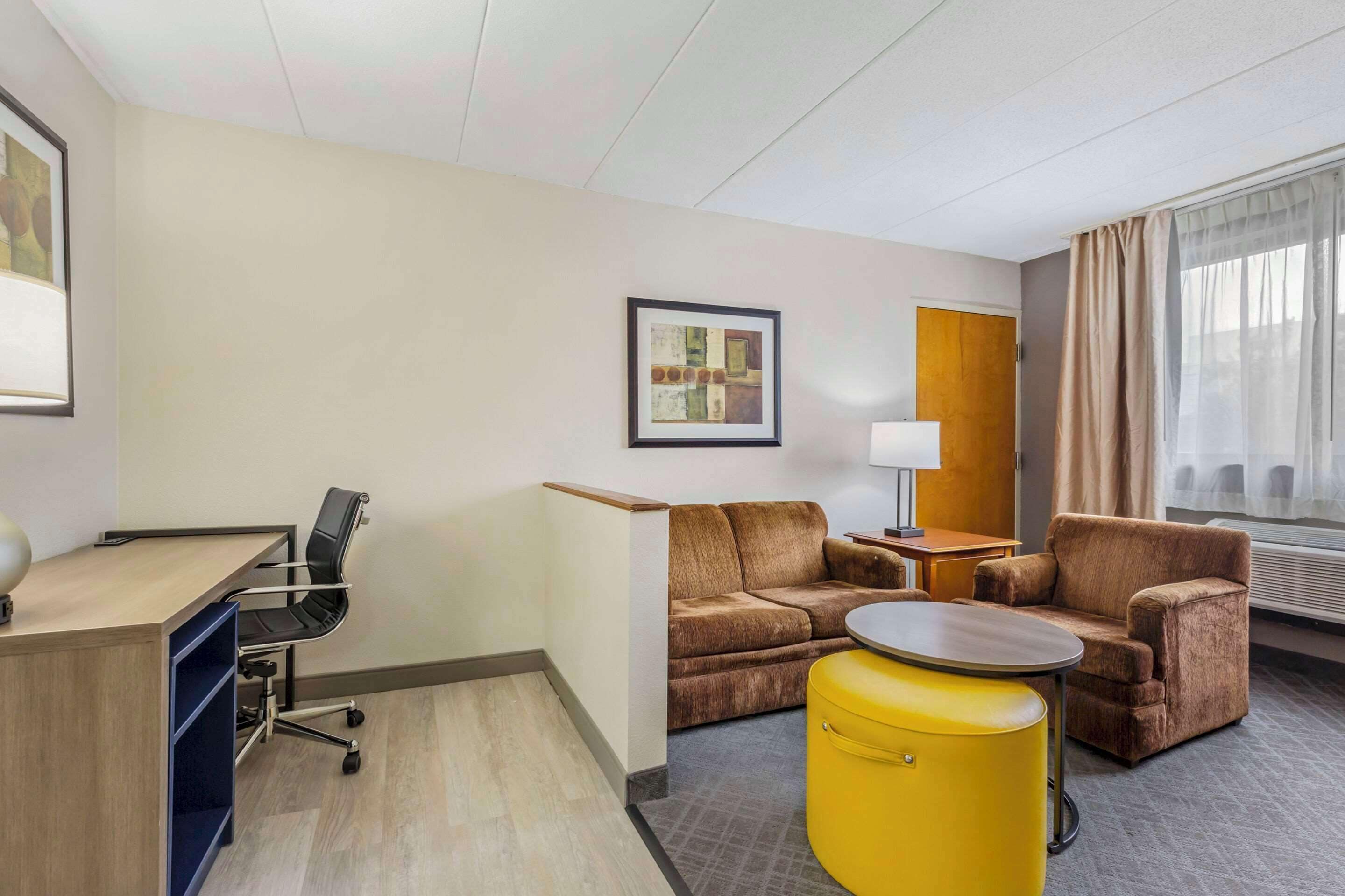 Une chambre d’hôtel équipée d’un bureau, de deux canapés, d’un pouf jaune, d’une lampe et d’un tableau encadré au mur.