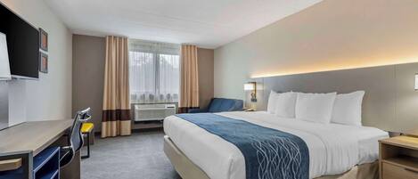 A hotel room with a large bed, a desk with a chair, a TV mounted on the wall, and a window with curtains.