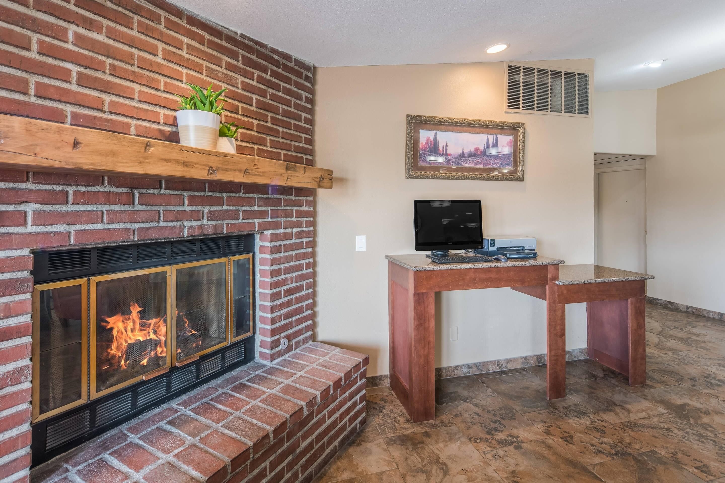 A fireplace with a brick surround, a wooden shelf with plants, a framed picture on the wall, and a desk with a computer.