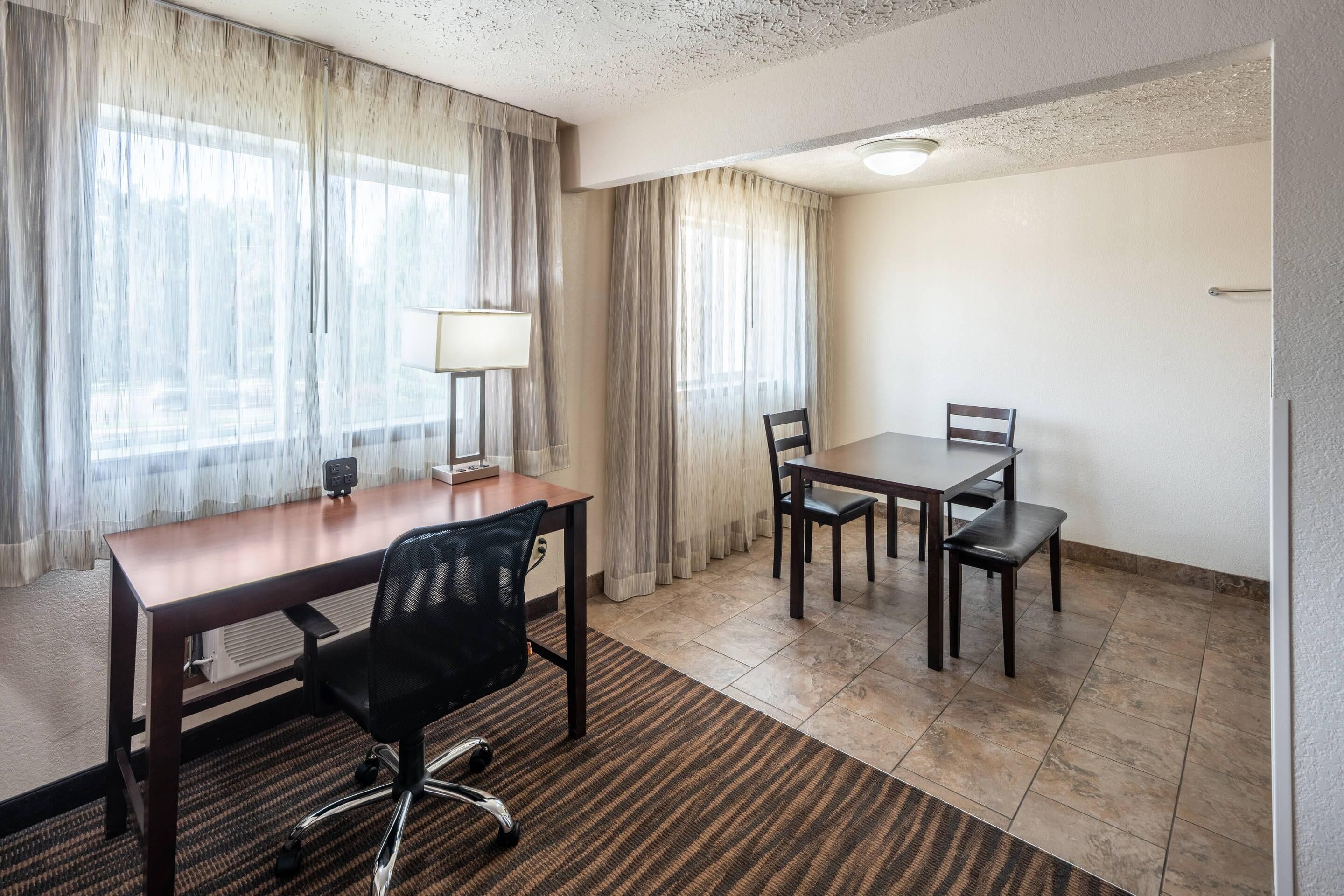 Una habitación de hotel con un escritorio de madera, dos sillas, una lámpara y un ventanal con cortinas.