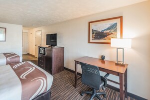 Una habitación de hotel con cama, escritorio, silla, televisor, lámpara y un cuadro en la pared.