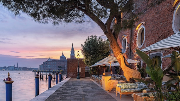 Une promenade en bord de mer avec des espaces de repos en plein air, un grand arbre et une vue sur un bâtiment historique situé de l’autre côté de l’eau.