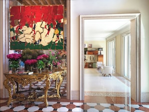A hallway with a gold ornate table, vibrant artwork, and a view into a room with a bookshelf and chairs.