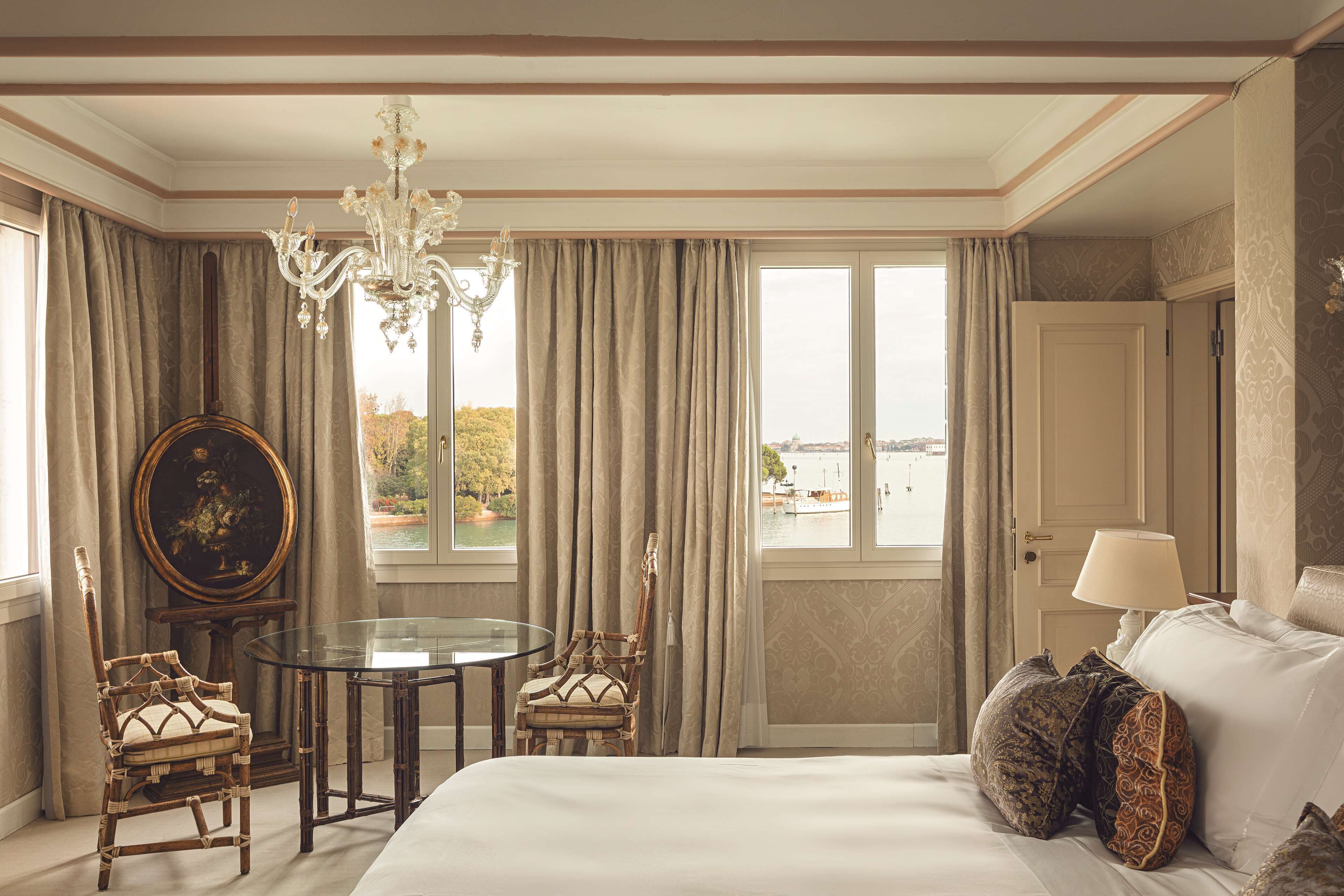 Une chambre d’hôtel avec un grand lit, une table ronde, deux chaises, un lustre et une vue sur une étendue d’eau par les fenêtres.