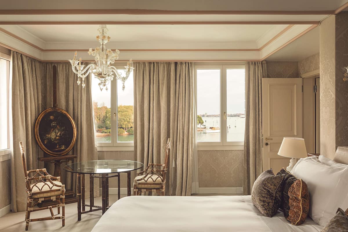 A hotel room with a large bed, a round table, two chairs, a chandelier, and a view of a body of water through the windows.
