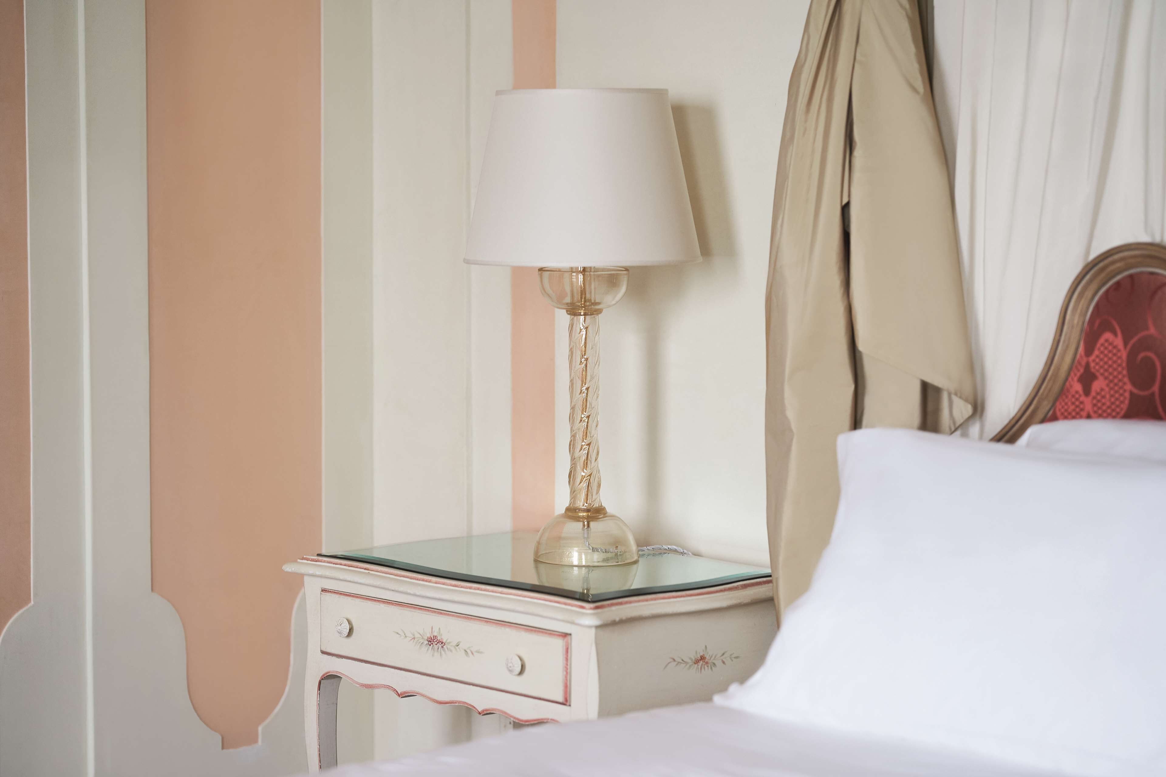 Un lit bien fait, avec du linge de lit blanc, une lampe de chevet à abat-jour blanc et une table de chevet en verre avec un tiroir.