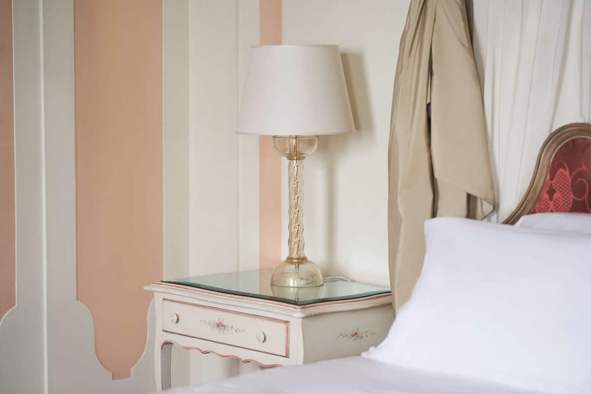 A neatly made bed with white linens, a bedside lamp with a white shade, and a glass-topped nightstand with a drawer.