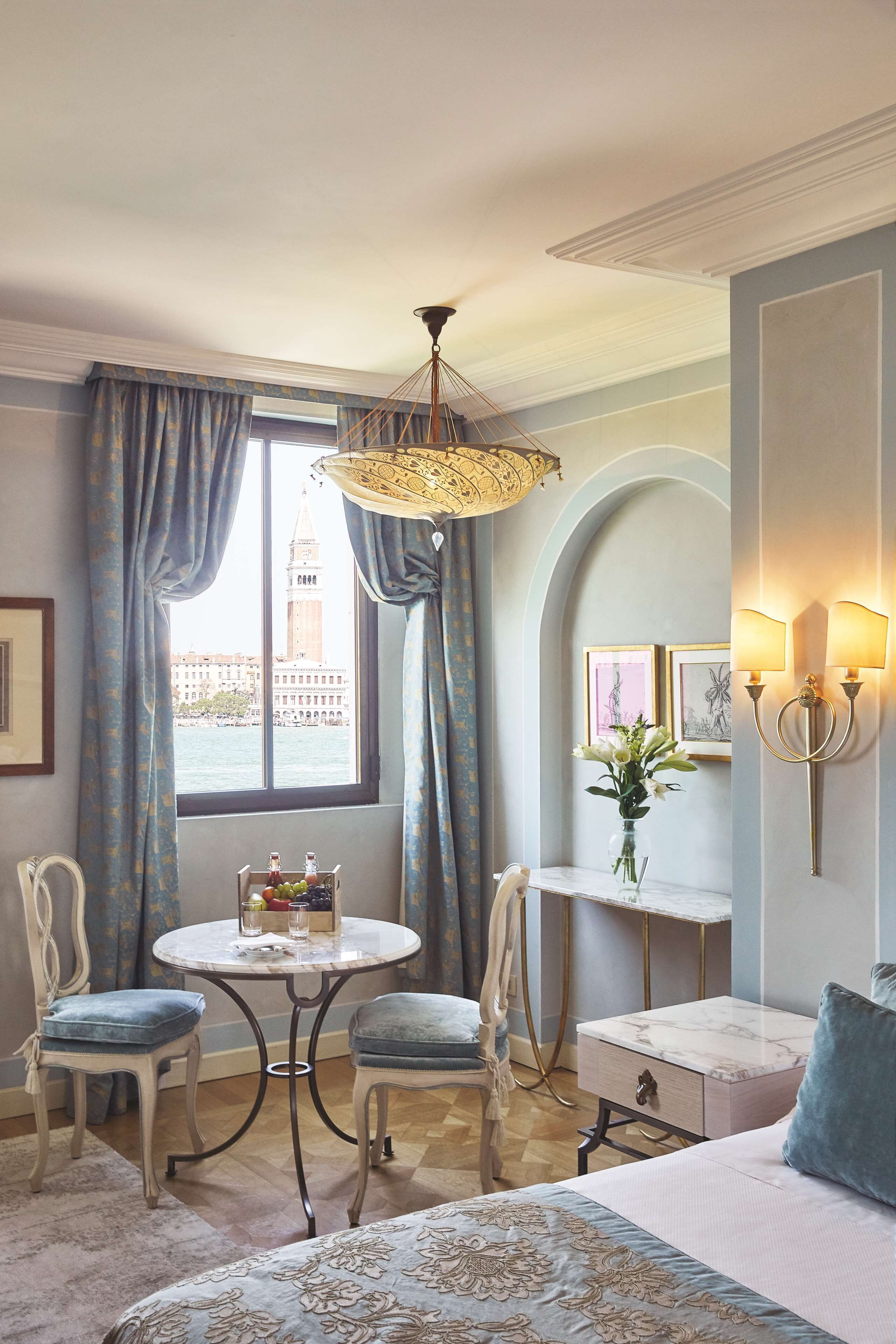 Une chambre d’hôtel avec un lit, une table ronde entourée de chaises, un vase de fleurs et une vue sur une tour par la fenêtre.