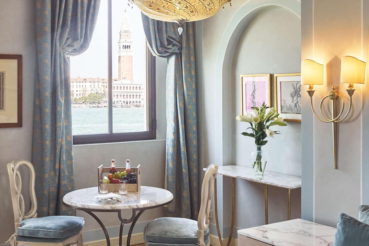 A hotel room with a bed, a round table with chairs, a vase with flowers, and a view of a tower through the window.
