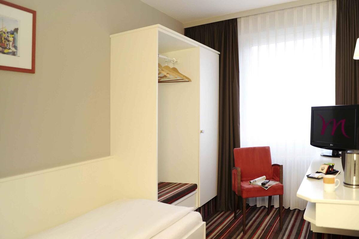 A hotel room with a bed, a red chair, a TV, a coat rack, and a window with curtains.