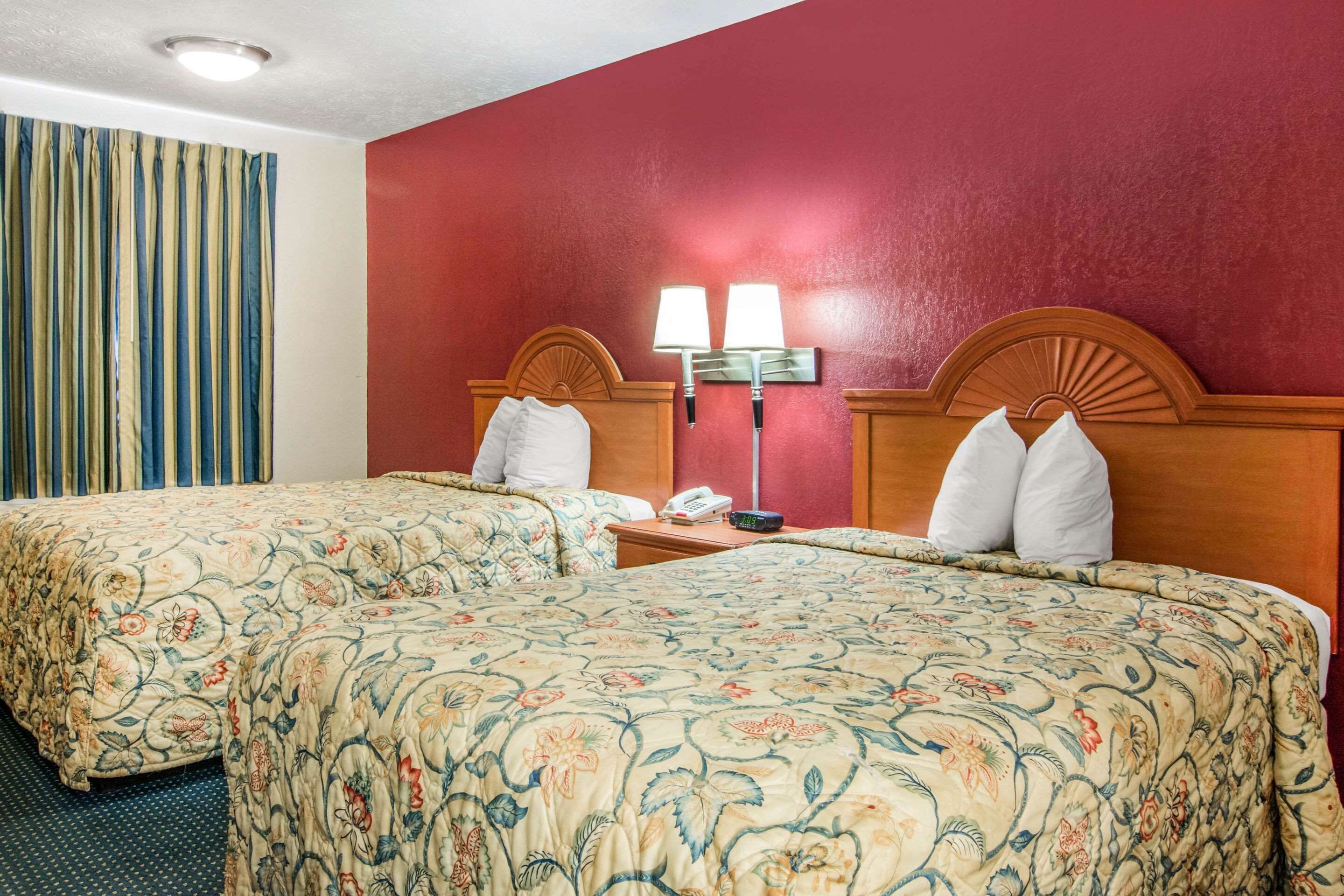 Habitación de hotel con dos camas, pared roja, cortina a rayas y mesita de noche con lámpara y teléfono.