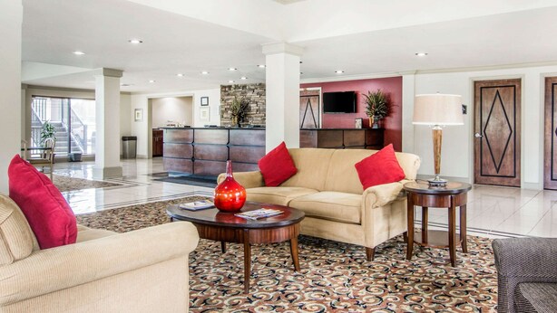 A spacious lobby with a beige sofa, red pillows, a coffee table, and a patterned rug. There is a stone wall, a television, and a door with a geometric design. - Rodeway Inn La Grange East (LaGrange)