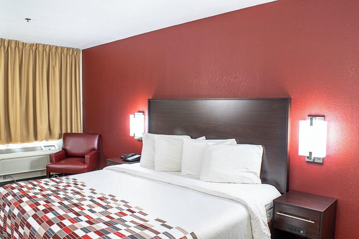 Una camera d'albergo con un letto grande, una poltrona rossa, un comodino con il telefono e una lampada a parete.