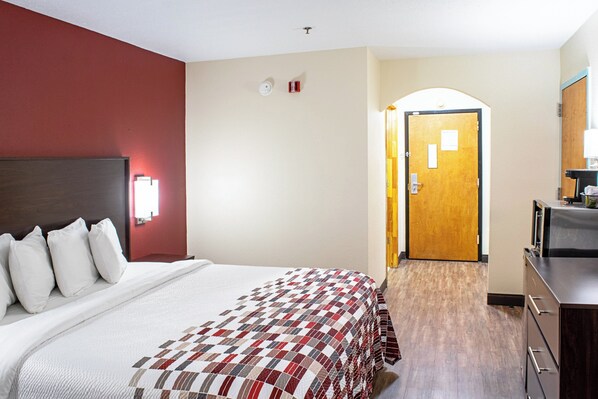 A hotel room with a large bed, a wooden headboard, a red accent wall, a door with a mirror, and a small desk with a lamp.
