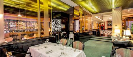 Un restaurant avec des tables dressées pour le service, un bar avec des bouteilles exposées et un sol recouvert d’une moquette verte.