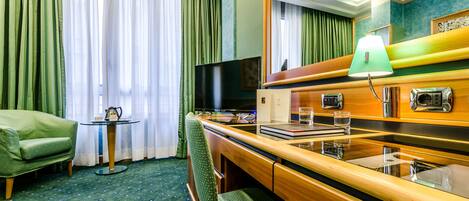 A hotel room with a green sofa, a small round table, a desk with a lamp, a television, and a mirror.