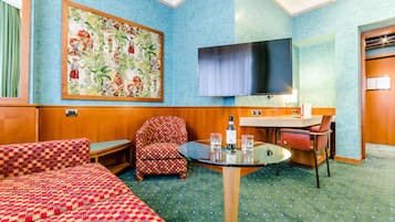 A hotel room with a patterned sofa, a small table with glasses, a television, and a large floral painting on the wall.