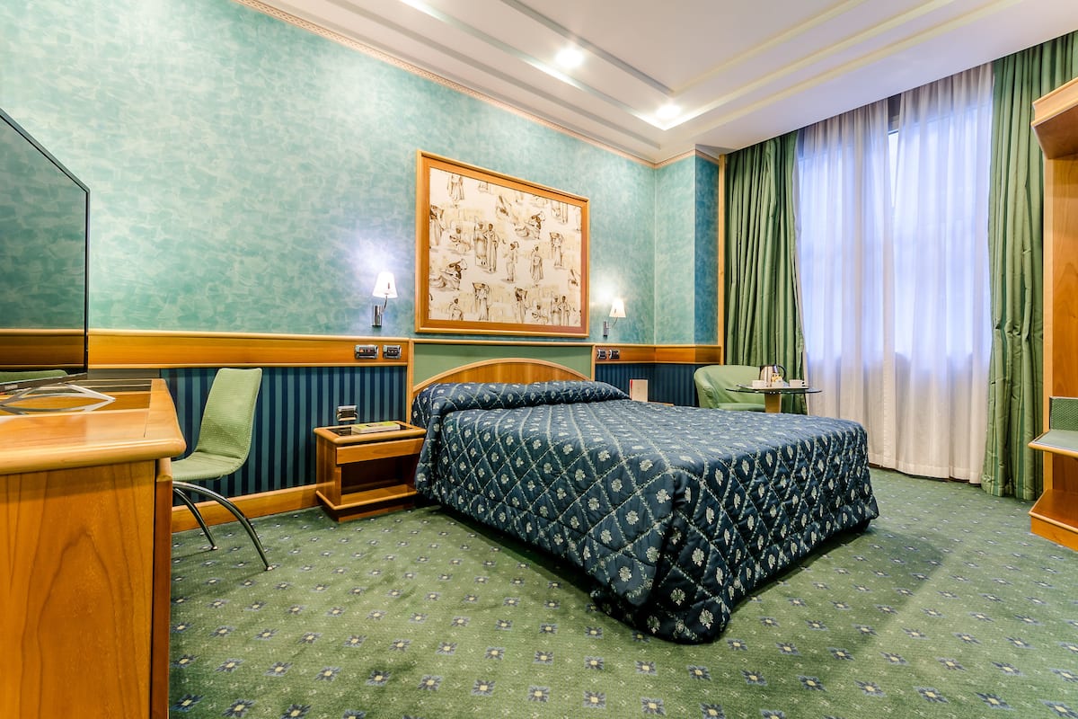 A hotel room with a large bed, a desk, a chair, a small table, and a painting on the wall.