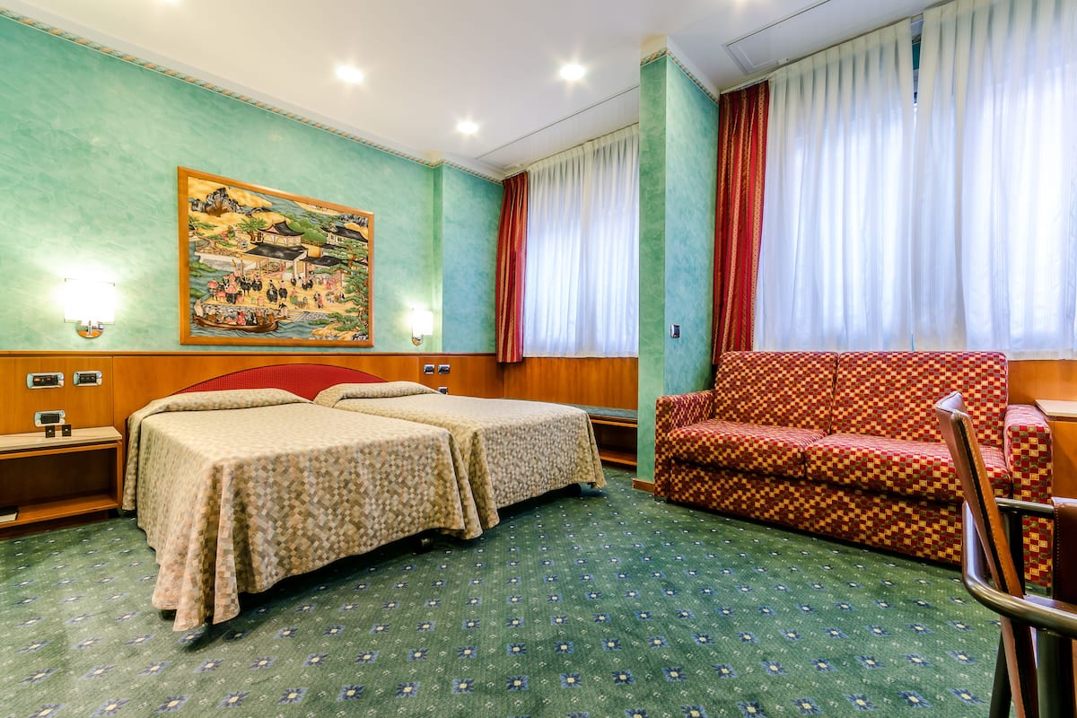A hotel room with two beds, a sofa, a painting on the wall, and a window with curtains.