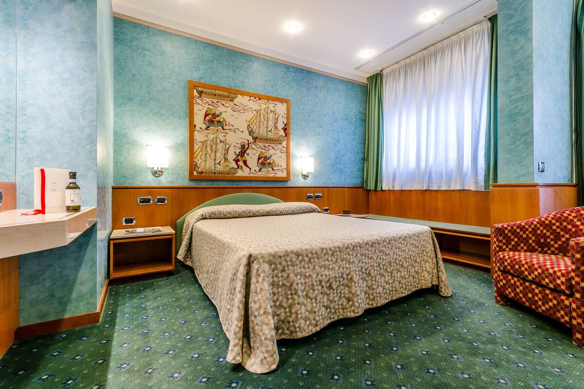 A hotel room with a bed, a chair, a nightstand, a lamp, and a wall map.