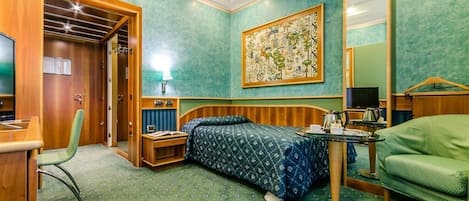 A hotel room with a bed, a desk, a chair, a table with a kettle, and a large mirror.