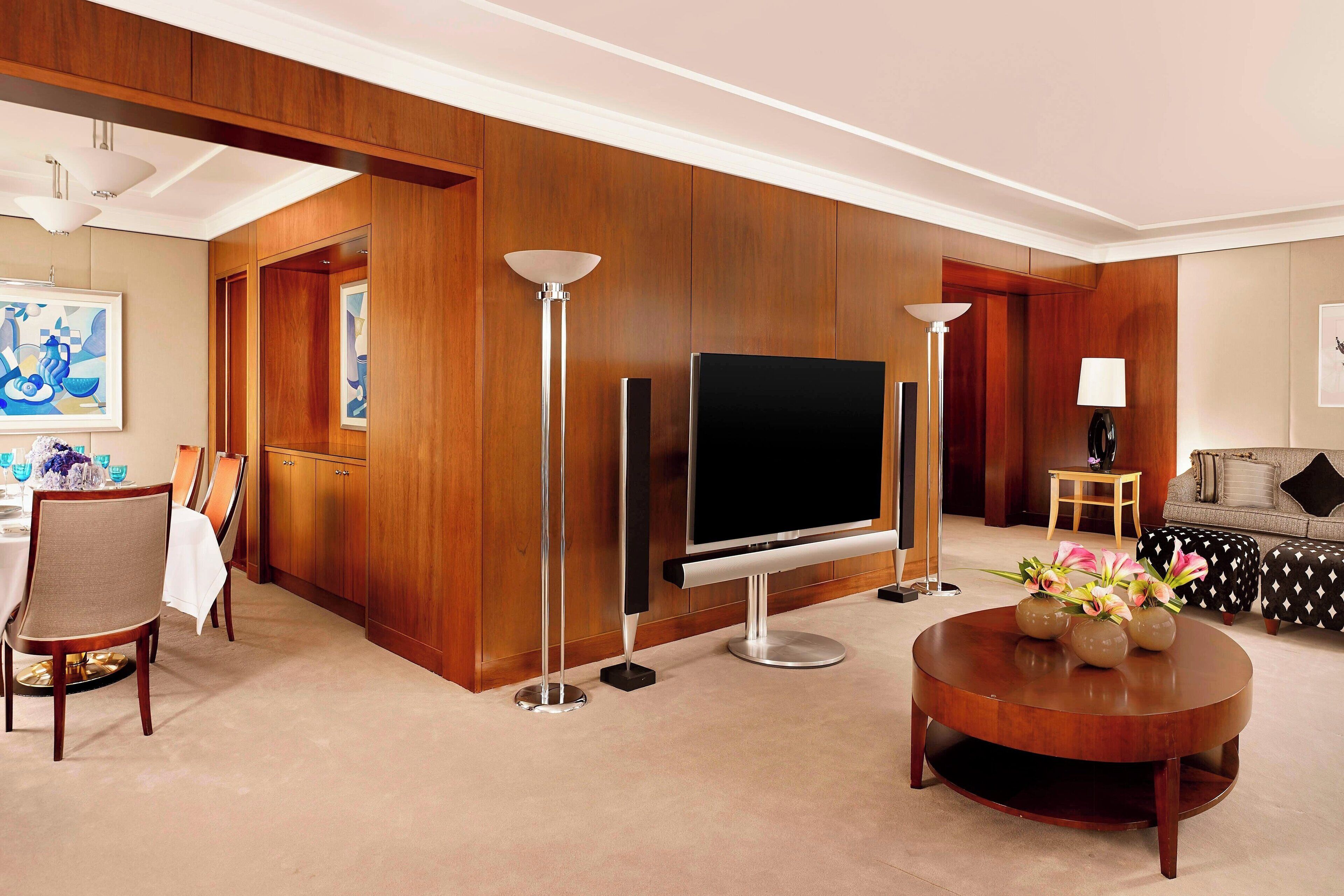 Una sala de estar moderna con televisor de pantalla plana, paneles de pared de madera, una mesa redonda con flores y un comedor con sillas y mesa.