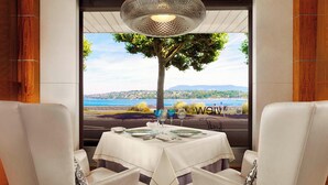 Un comedor con una mesa dispuesta para dos personas, vista a un árbol y paisaje a través de una ventana, y una lámpara decorativa en el techo.