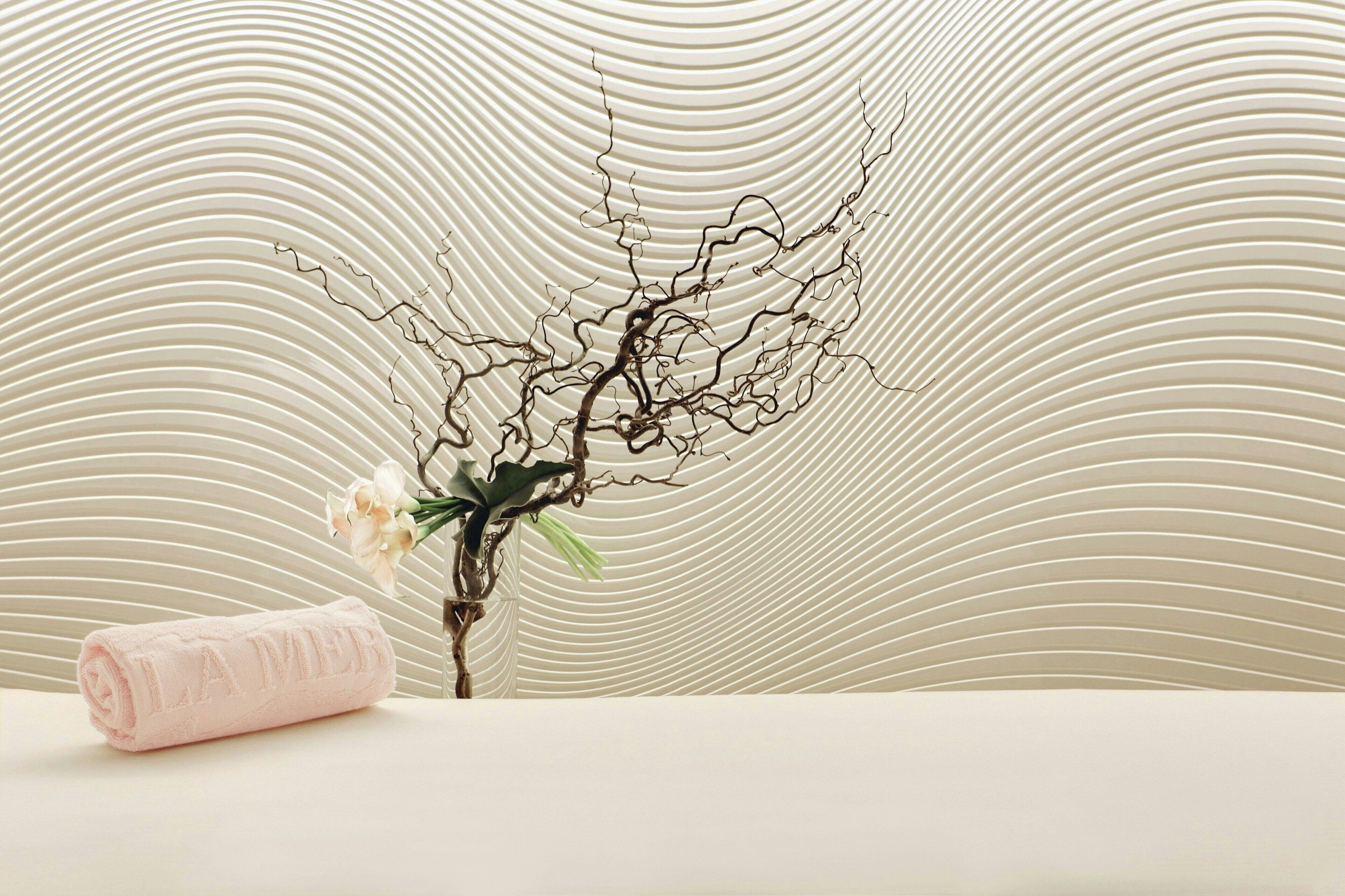 Une serviette soigneusement enroulée, portant en relief l’inscription « LA MER », une branche ornée de fleurs et, en arrière-plan, un mur à texture.