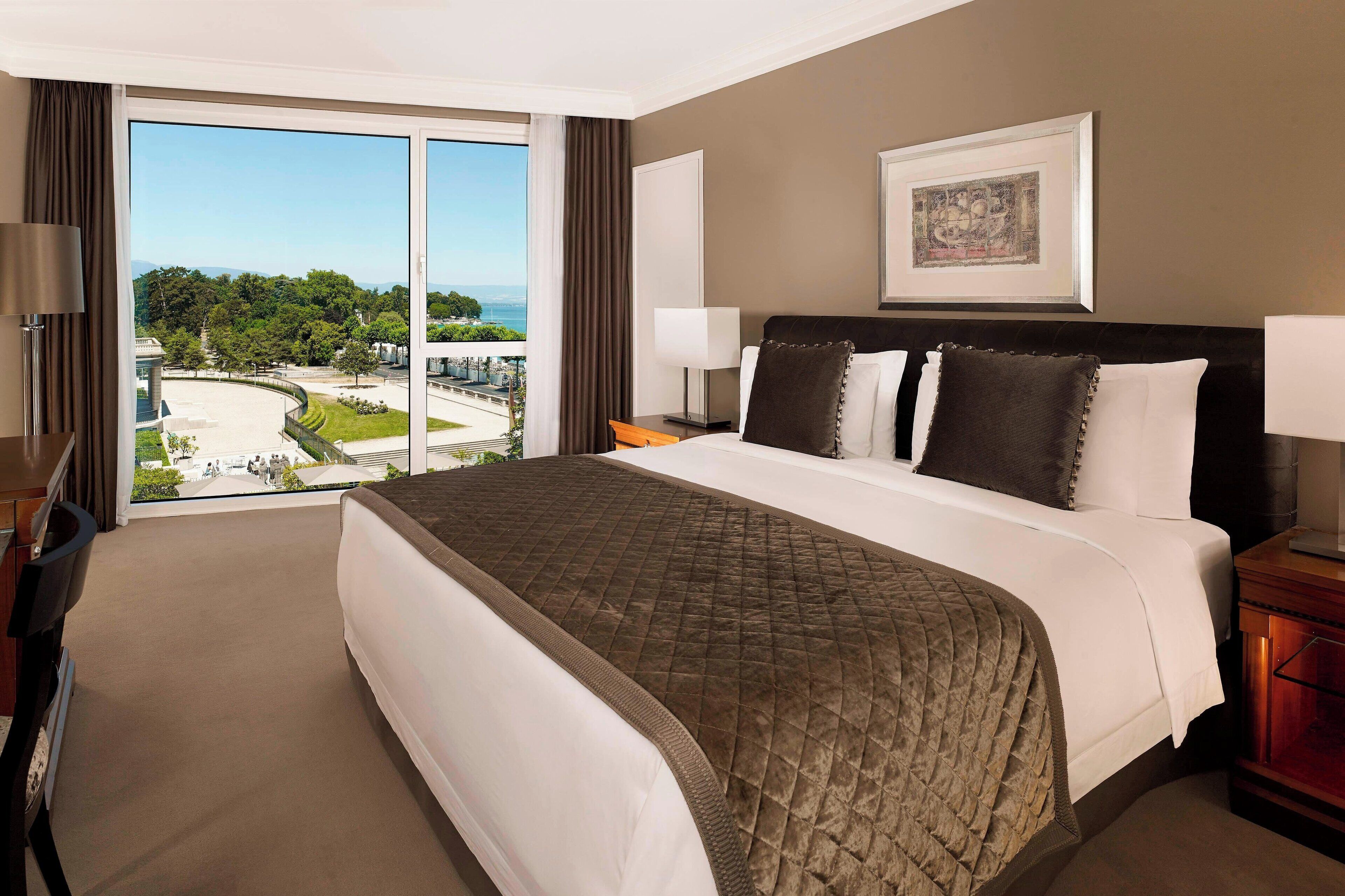 A hotel room with a large bed, a view of a cityscape, and a framed picture on the wall.
