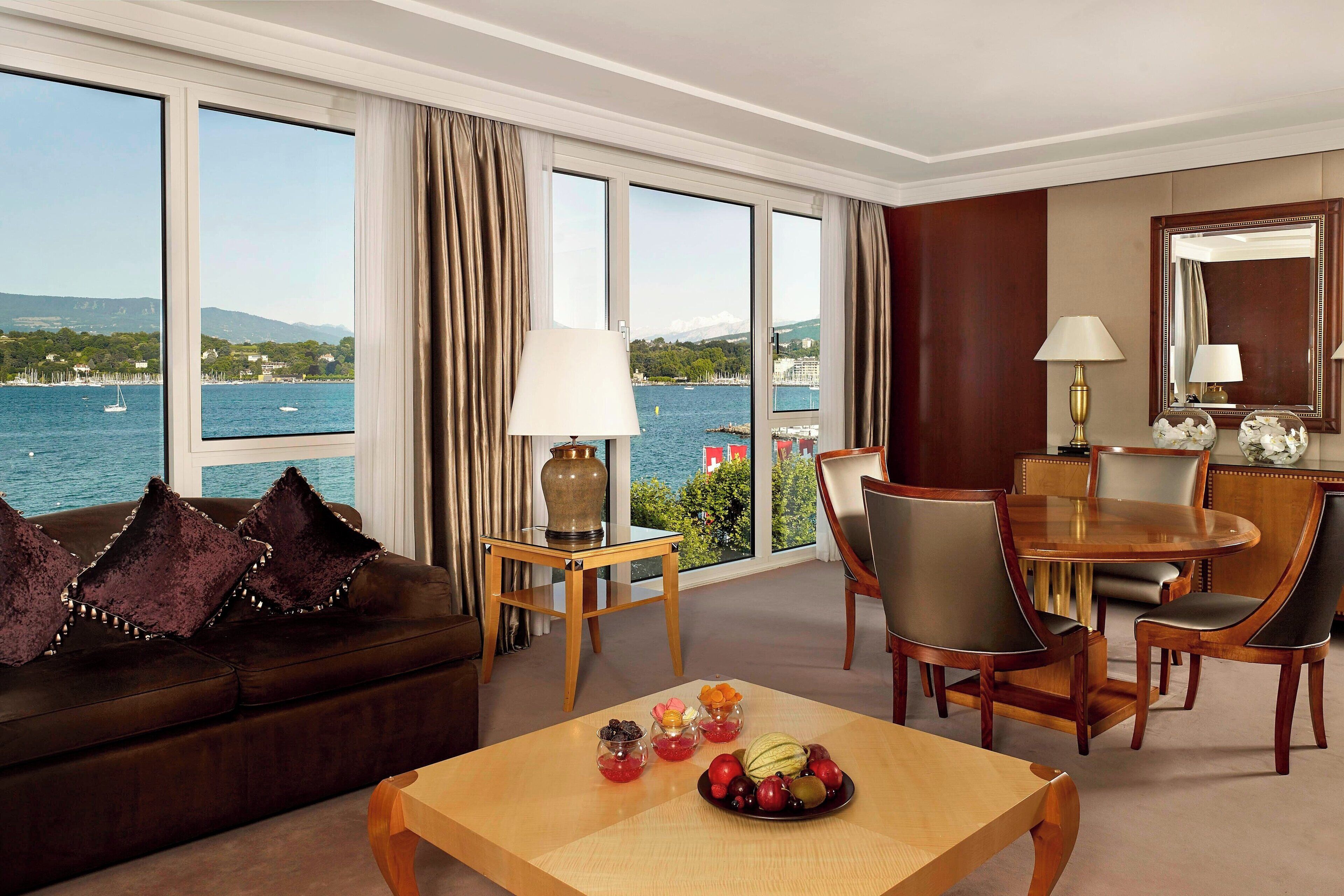 A hotel room with a large window offering a view of the sea and mountains, a wooden dining table with chairs, a sofa with decorative pillows, and a small side table with a lamp.