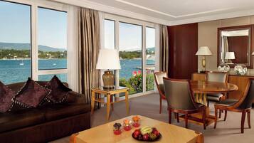 A hotel room with a large window offering a view of the sea and mountains, a wooden dining table with chairs, a sofa with decorative pillows, and a small side table with a lamp.