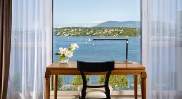 Ein Holztisch mit einer Vase voller weißer Blumen, ein Stuhl und Blick auf einen See mit Segelbooten und einer Stadt am Hang.