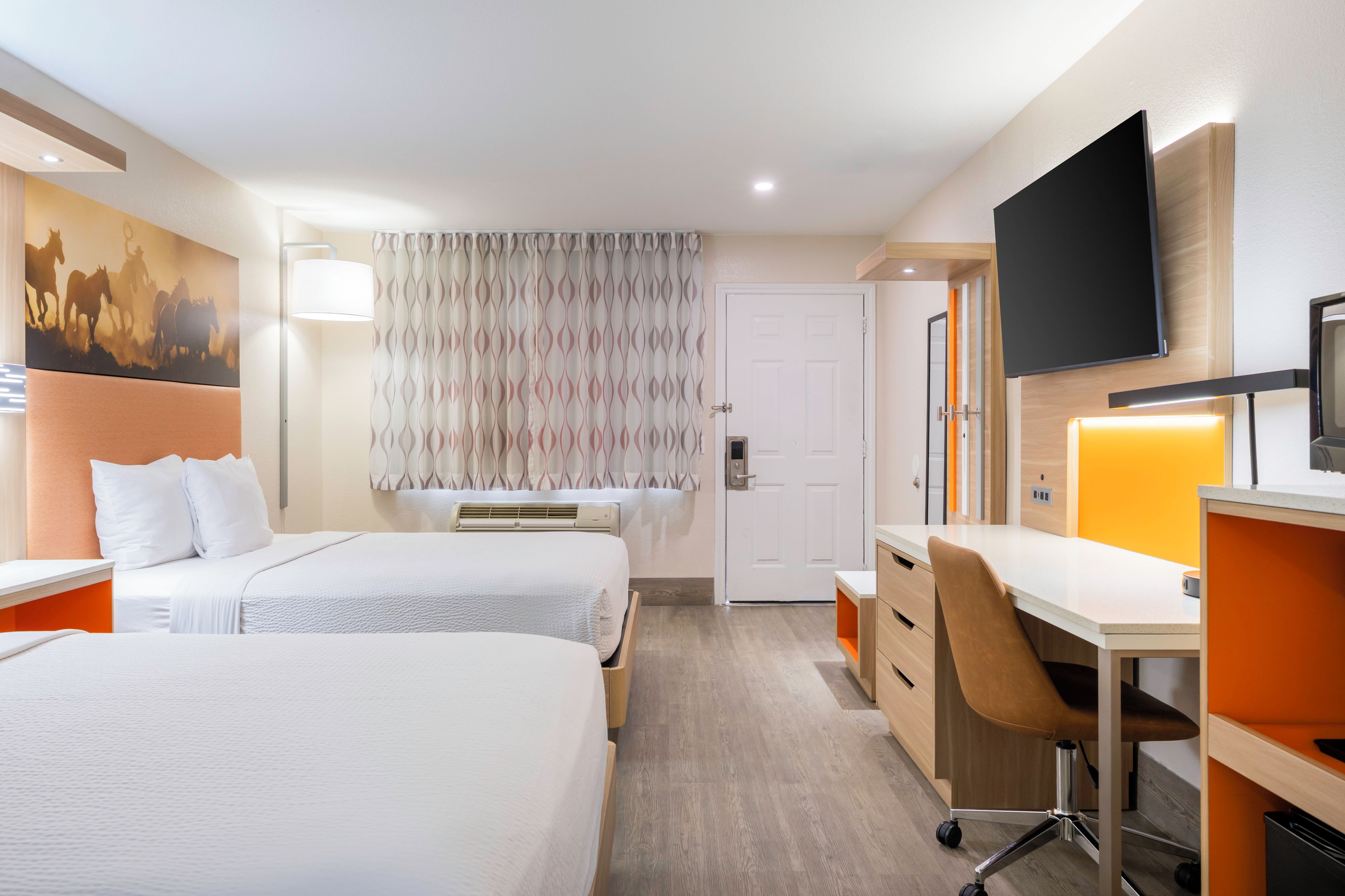 Quarto de hotel com duas camas, uma escrivaninha, uma cadeira, uma televisão e um quadro na parede.
