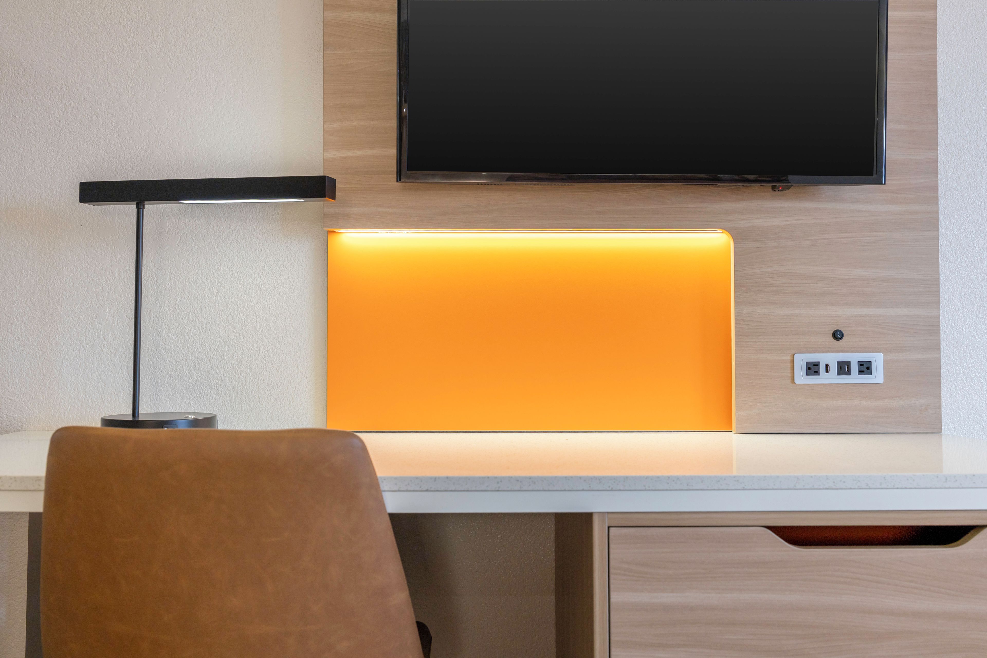 Habitación de hotel con televisión de pantalla plana montada en la pared, un escritorio con lámpara y una silla.